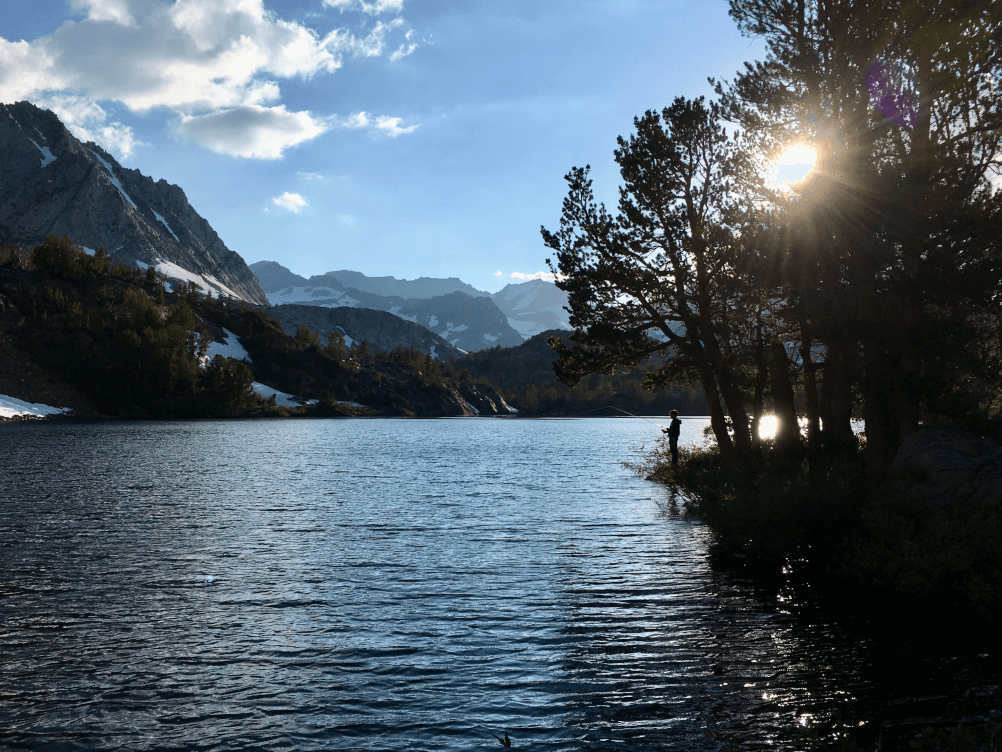 Solitary angler fishing at sunset in alpine lake with snow capped peaks in the background. In the high Eastern Sierra.